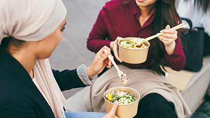 Dos mujeres comparten una comida, comen con cubiertos de plástico y un cuenco de cartón.